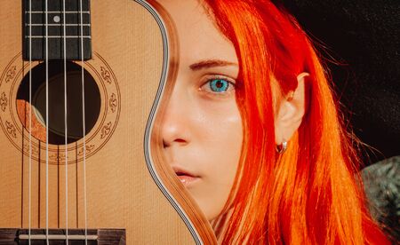 Sexy redhead girl with long hair plays the ukulele. Perfect woman portrait. Gorgeous strong and thick hair. Deep blue eyes. Natural beauty, clean skin, face and hair careの写真素材
