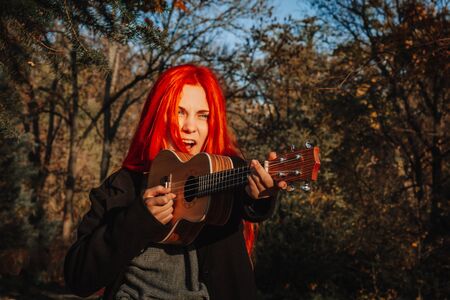 Red-haired girl with long hair plays on the ukulele in the park. School, music education concept, student learns to play the string instrument. Hands of a musician, classical, melody, creativityの写真素材