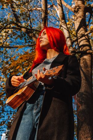 Red-haired girl with long hair plays on the ukulele in the park. School, music education concept, student learns to play the string instrument. Hands of a musician, classical, melody, creativityの写真素材