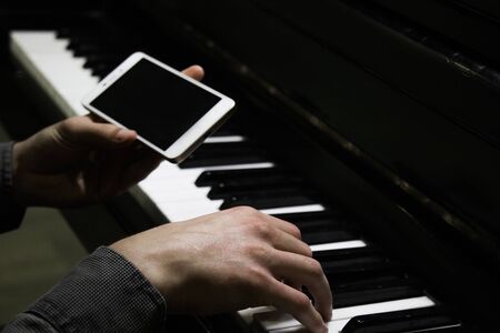 two male hands on the piano with a smartphone. palms lie on the keys and play the keyboard instrument in a music school. student learns to play. hands of a pianist. black dark backgroundの写真素材