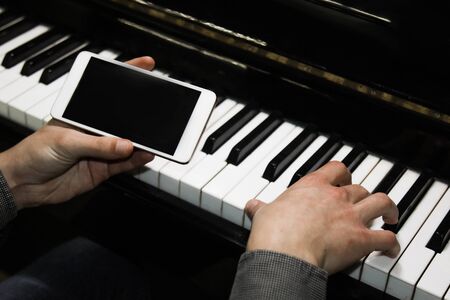 two male hands on the piano with a smartphone. palms lie on the keys and play the keyboard instrument in a music school. student learns to play. hands of a pianist. black dark backgroundの写真素材