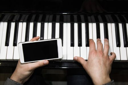 two male hands on the piano with a smartphone. palms lie on the keys and play the keyboard instrument in a music school. student learns to play. hands of a pianist. black dark backgroundの写真素材
