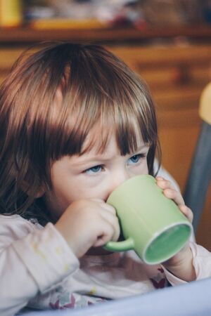 A one-year-old smiling girl sits at a children's table in a high chair and eats with a spoon from a bowl. Colored background. Healthy eating for kids. Baby foodの写真素材