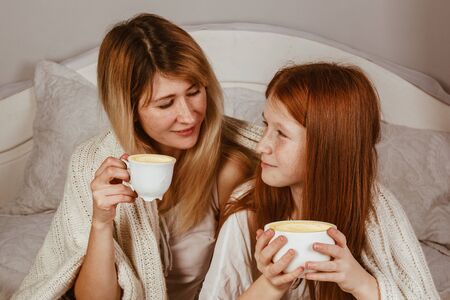 Mom and ginger-haired daughter are sitting on the bed and drinking cocoa with froth. Covered with a blanket, hugの写真素材