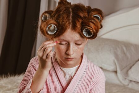 Young beautiful girl does makeup on the bed in the bedroom. Red-haired and caulking with curlers on his head in a bathrobeの写真素材