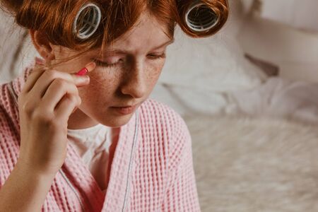 Young beautiful girl does makeup on the bed in the bedroom. Red-haired and caulking with curlers on his head in a bathrobeの写真素材