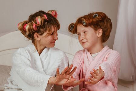 Happy loving family. Mom and daughter do manicures, pedicures, do makeup and have fun. Mom and little girl in bathrobes and with curlers on their headsの写真素材