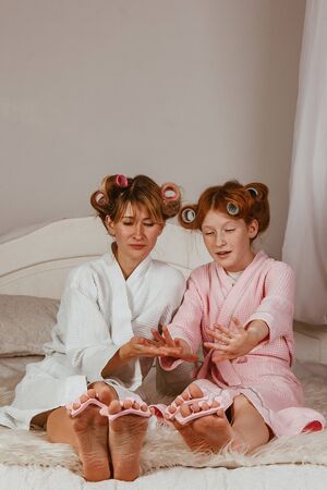 Happy loving family. Mom and daughter do manicures, pedicures, do makeup and have fun. Mom and little girl in bathrobes and with curlers on their headsの写真素材