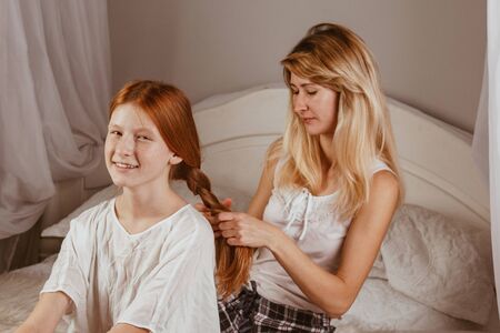 Happy family - a smiling mother braids her daughter's hair in the morning after sleeping in bed. Mom gathers daughter to school. Interaction, contact, loveの写真素材