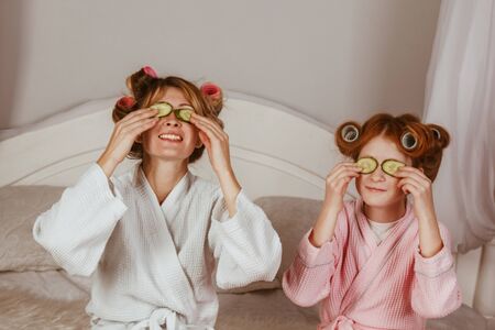Holiday day at home. Mom and daughter at home make a mask of cucumber on the eyes. Beauty Salon Concept. Beautiful face. Woman and happy baby. Girls with curlersの写真素材
