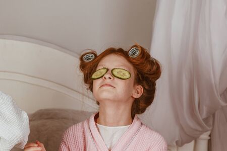 Holiday day at home. Mom and daughter at home make a mask of cucumber on the eyes. Beauty Salon Concept. Beautiful face. Woman and happy baby. Girls with curlersの写真素材