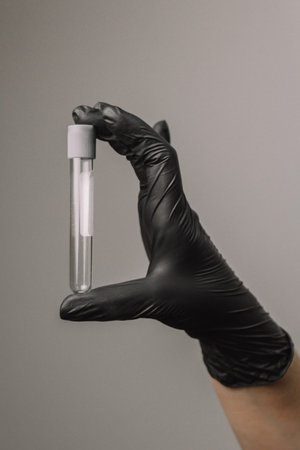 Empty test tube in a glass flask in the hands of a scientist or cosmetologist. Black gloves on a gray background. Experiment, analyzes, girl from the backの写真素材