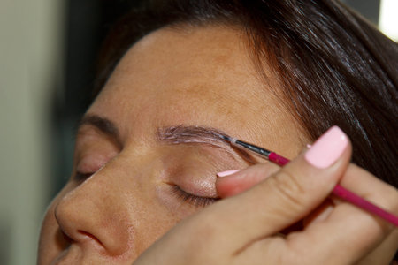 cosmetologist - makeup artist paints henna on previously plucked, design, trimmed eyebrows in a...