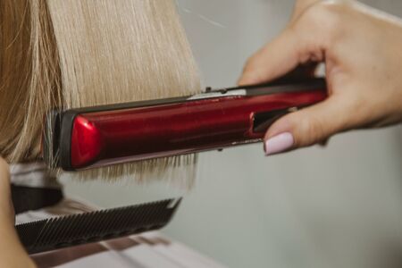 Hairstyle. Attractive blonde with short hair does a hairstyle with an electric iron. Hair straightener gray background. close-up. Macro photoの写真素材