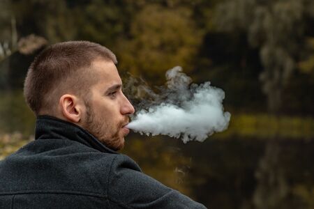 A guy with a beard sits on the lake and smokes an electronic cigarette. Blows puffs of smoke. The concept of unity with nature. Rest near the water. Vape, Man Vaping. lips and smoke, a lot of smokeの写真素材