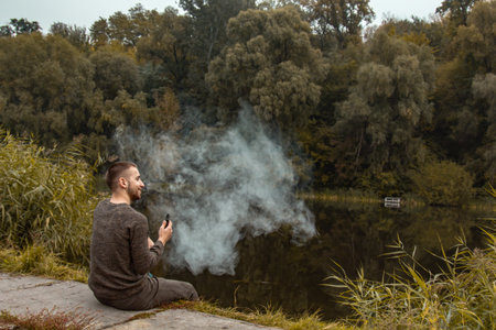 A guy with a beard sits on the lake and smokes an electronic cigarette. Blows puffs of smoke. The...