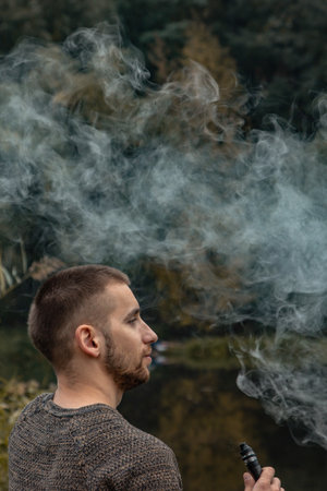 A guy with a beard sits on the lake and smokes an electronic cigarette. Blows puffs of smoke. The...