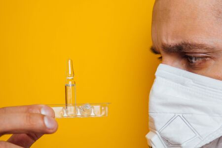 Portrait of a young doctor in a respirator on a yellow background. Holding an ampoule with a coronavirus vaccine in his handsの写真素材