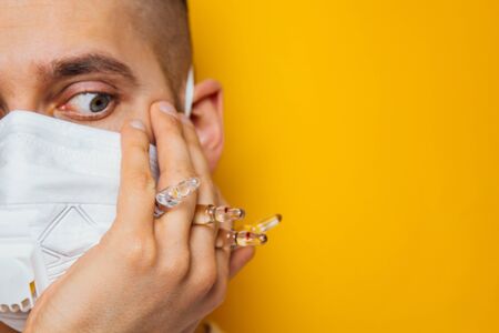 Portrait of a young doctor in a respirator on a yellow background. Ampoules with a coronavirus vaccine in the hand. Look into the camera. The inventor of the drug. The concept of dry diseaseの写真素材