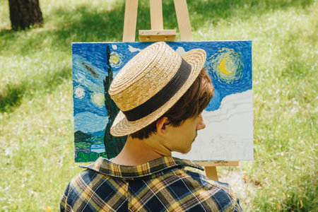 a young guy in a straw hat sits in the park in front of an easel and paints a picture with oil...