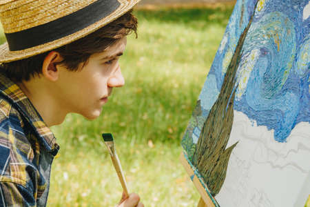a young guy in a straw hat sits in the park in front of an easel and paints a picture with oil paints. Artistic work in nature. Plein air.の写真素材