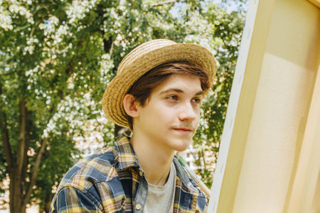 a young guy in a straw hat sits in the park in front of an easel and paints a picture with oil...
