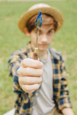a young guy in a straw hat sits in the park in front of an easel and paints a picture with oil paints. Artistic work in nature. Plein air.の写真素材