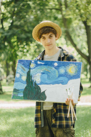 a young guy in a straw hat sits in the park in front of an easel and paints a picture with oil...