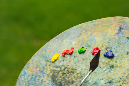 the hand of a European artist holds a mastekhin smeared in oil paint and a palette against the background of a green lawn. Blurred background. copy space. Mixing board.の写真素材