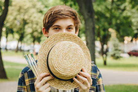 a young guy with brushes for painting stands in the park against the background of trees in summer on a sunny day. Straw Hat Artist.の写真素材