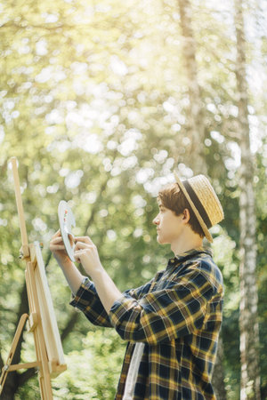 a young guy in a straw hat sits in the park in front of an easel and paints a picture with oil paints. Artistic work in nature. Plein air.の写真素材
