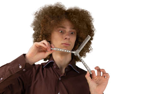 A long-haired curly-haired guy in a brown shirt on a white background uses a metal comb. Emotions before a haircut in a hairdresser.の写真素材