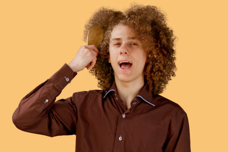 A long-haired curly-haired guy in a brown shirt on an orange background uses a wooden comb. Emotions...