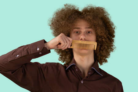 A long-haired curly-haired guy in a brown shirt on a blue background uses a wooden comb. Emotions before a haircut in a hairdresser.の写真素材