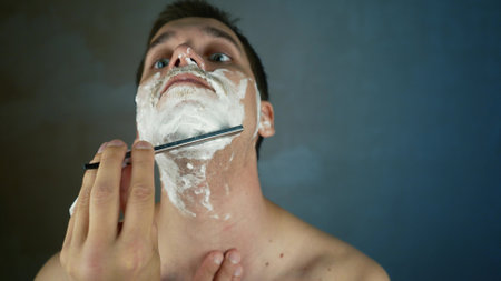 A young handsome guy shaves his beard with a dangerous razor. Trimming the beard with an old-fashioned blade in the bathroom. Handsome macho man shaved his beard at home. close-up shotの写真素材