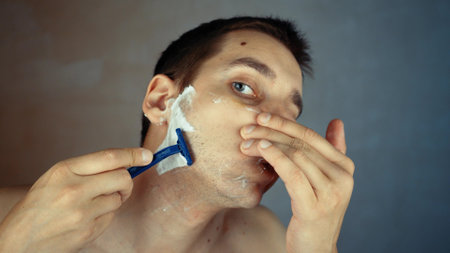 Young man is shaving using disposable razor in bathroom, closeup. Daily morning routine, hygienic procedure. Man is shaving beard and mustacheの写真素材