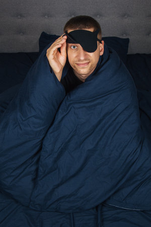 Embarrassed young man in a blanket at home wears a sleep mask while relaxing at home, studio portrait. Relax, lifestyle concept in a good moodの写真素材