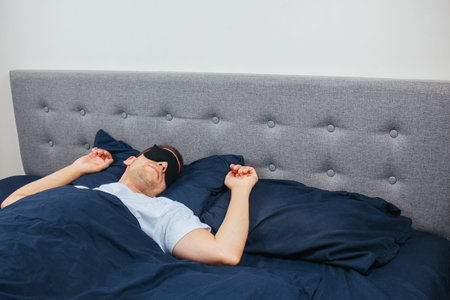 A man wrapped in a blanket wearing a sleep mask on a gray background. Sleeping at home in his bed....