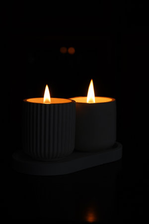 A set of lit white candles flickering in the dark on a black background with one candle in focus in the foreground. Lighting a candle. A burning candle. Beautiful handmade candles. Cozy atmosphere in the houseの写真素材