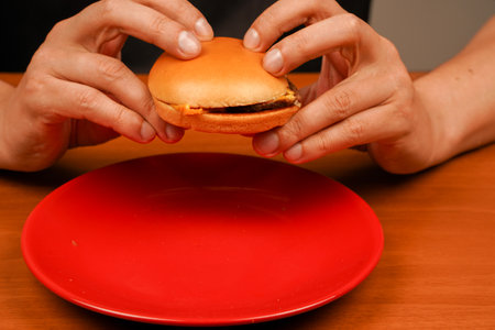 Close up shot of young man eats burger. Man holds burger with hands. Hands holding homemade beef burger on wooden table. Ready to eat tasty fast food hamburger.の写真素材