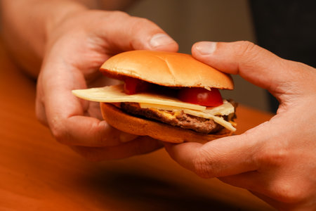 Close up shot of young man eating burger. Man holds burger with hands. Hands holding homemade beef burger on wooden table. Ready to eat tasty fast food hamburger.の写真素材
