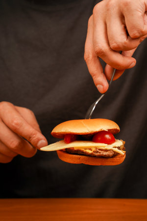 Close up shot of young man eats burger. Cutting homemade beef burger on wooden table. Ready to eat tasty fast food hamburger. Unhealthy food concept. Man holds burger with hands. Home delivery.の写真素材
