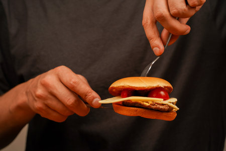 Man holds burger with hands. Man eats beef burger on wooden table. Ready to eat tasty fast food hamburger. Unhealthy food concept. Home delivery. Close up shot of young man eats burger.の写真素材