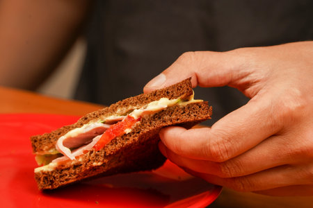Close-up of man eating sandwich. Close-up image of a male cooking and holding sandwich. Food. Tasty sandwich. Hands holding sandwich with ham, bacon, cheese, lettuce, tomatoes.の写真素材