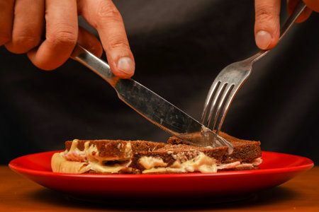 Close-up of man eating sandwich. Close-up image of a male cooking and holding sandwich. Food. Tasty sandwich. Cutting sandwich with fork and knife. Sandwich with ham, bacon, cheese, lettuce tomatoes.の写真素材