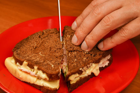 Close-up of man eating sandwich. Close-up image of a male cooking and holding sandwich. Food. Tasty sandwich. Cutting sandwich with fork and knife. Sandwich with ham, bacon, cheese, lettuce tomatoes.の写真素材