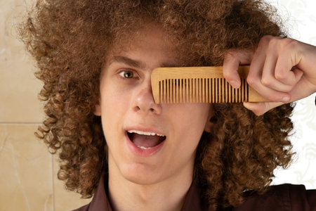 Hair care concept. Smiling curly haired young man holding brush closing one eye, studio shot.の写真素材