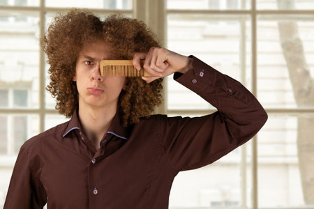 Looking at camera while taking care of his hair. Isolated window background. Smiling curly-haired bearded young man.の写真素材