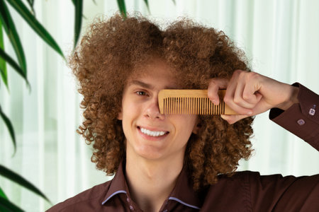 Smiling curly haired young man holding brush closing one eye, studio shot. Hair care concept.の写真素材