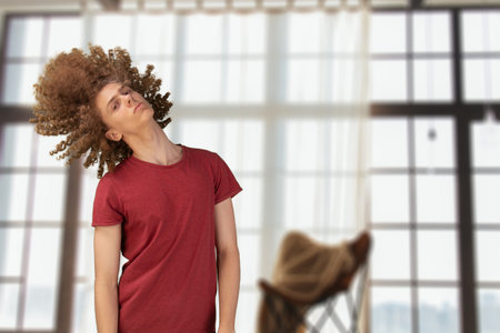 Man waving his curly hairdo. Young guy in dark red t-shirt struggling from tangled curly hair, standing in studio, copy space. Dry and matted hair.の写真素材